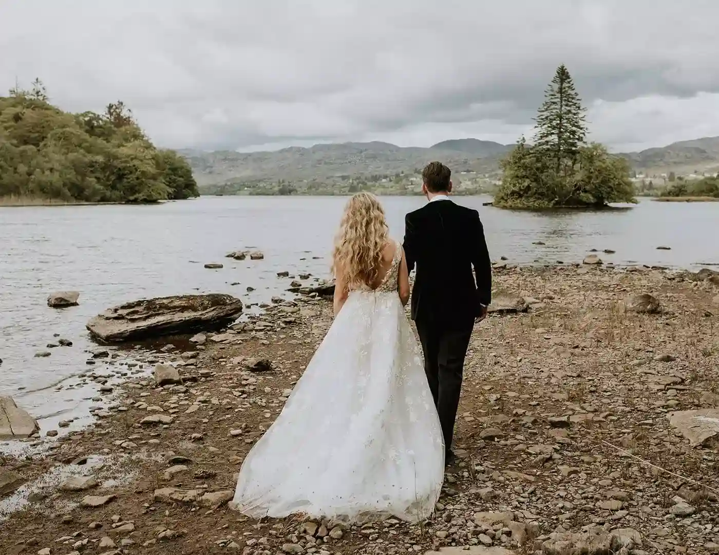 Bride in a white wedding gown and groom in a black suit stand hand in hand on a rocky lakeshore, facing the water and distant hills.