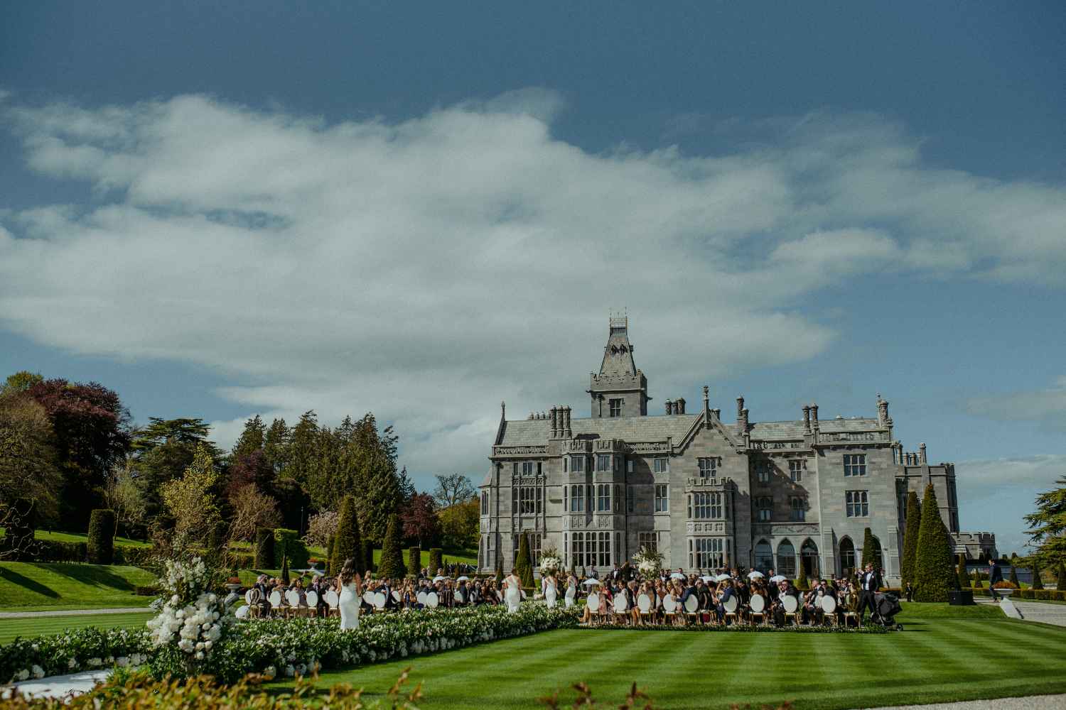 Outdoor wedding ceremony on the lawn of a gray stone manor, guests seated in rows of white chairs amid white floral arrangements.