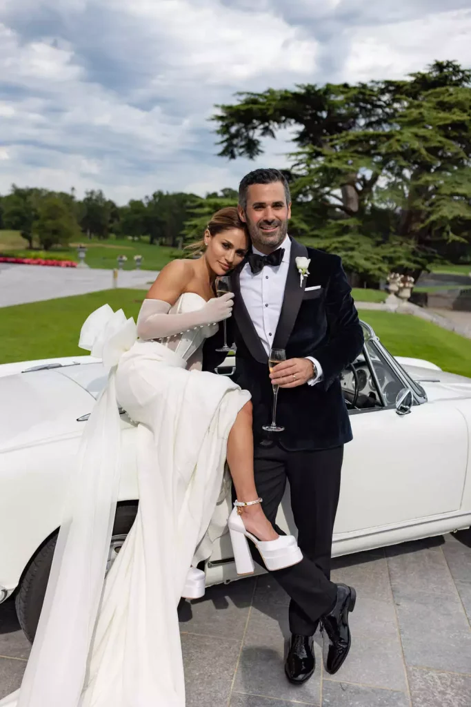 Bride, wearing a gown designed by Mark Ingram, and Groom, wearing blue velvet Alexander Nash suit, sit on vintage car outside Adare Manor