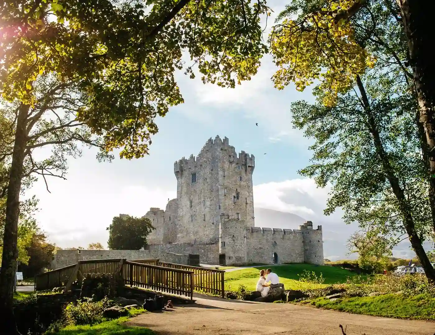 Ross Castle, County Kerry — couple seated beneath mature trees with the medieval castle rising behind them during an outdoor portrait session.