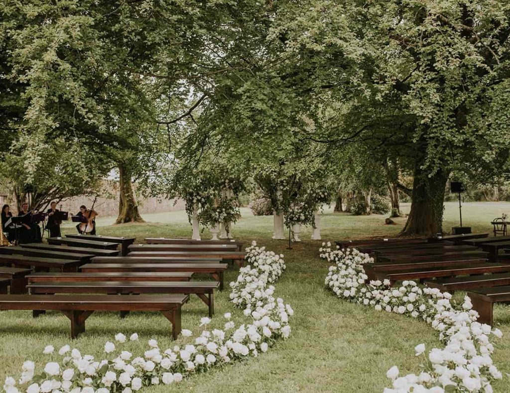 Castle wedding in Ireland at Lough Eske Castle, County Donegal — rose-lined forest aisle with wooden ceremony seating beneath mature trees on the estate grounds.