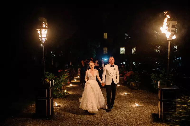 Castle weddings Ireland at Ballynahinch Castle, County Galway, Ireland — bride and groom leading guests to the late-night party while holding hands, illuminated by tall outdoor fire lamps along the evening pathway.
