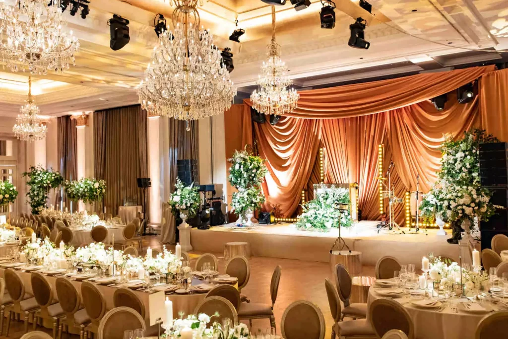 Destination wedding at Adare Manor, County Limerick — ballroom prepared for the dinner reception with banquet tables, a stage, chandeliers, and white floral arrangements with greenery.