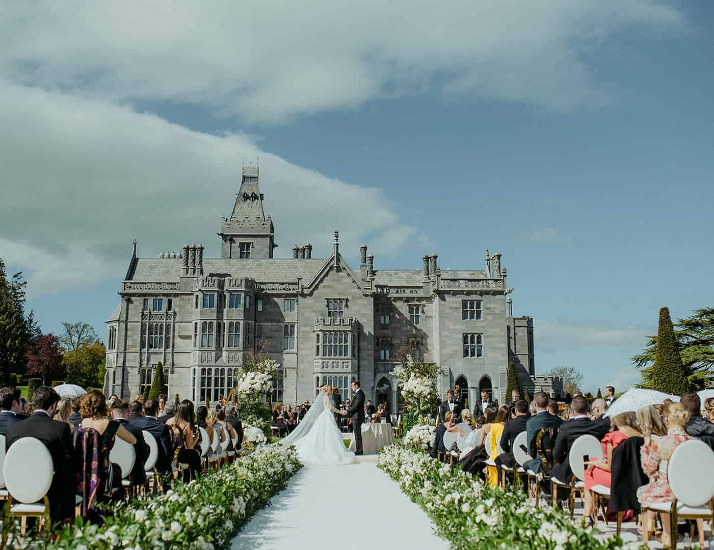 Destination wedding ceremony at Adare Manor, County Limerick — bride and groom holding hands at the altar during an outdoor ceremony, with guests seated along the aisle and the manor in the background.