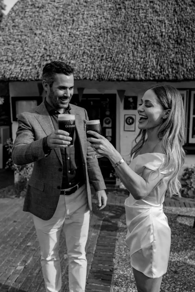 Destination wedding in Ireland at Adare Manor, County Limerick — bride and groom smiling during rehearsal dinner while holding pints of Guinness.