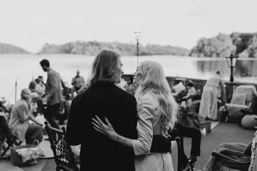 Couple with arms around each other at a lakeside outdoor party, facing away while guests mingle in the background.