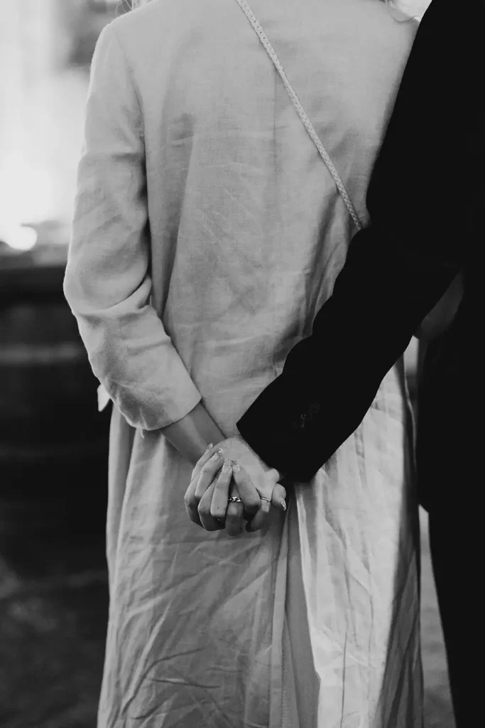 Two people standing side by side, holding hands behind their backs; one in a light dress and the other in a dark outfit with a crossbody strap.