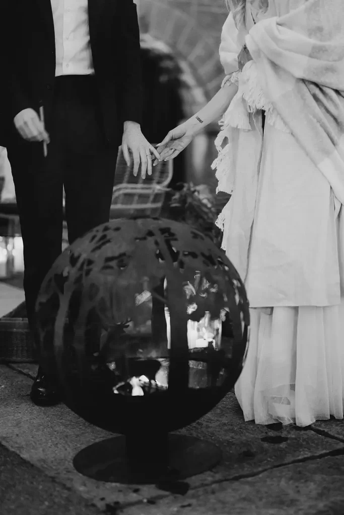 Destination wedding ceremony at Ashford Castle, County Mayo — circular bonfire feature in the foreground with the bride and groom in a candid moment behind, reaching to hold hands.
