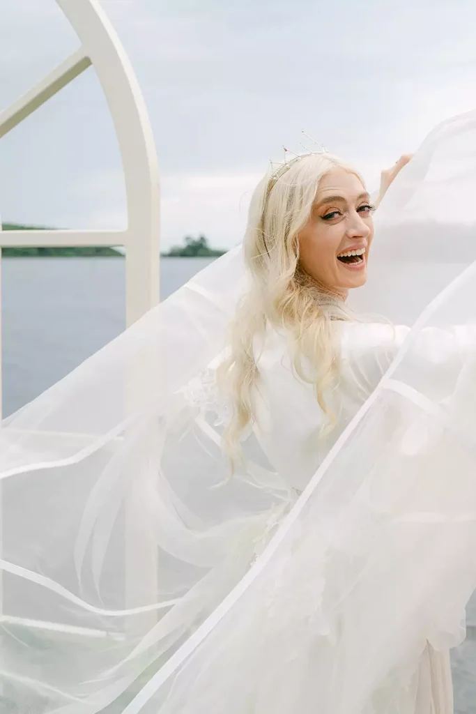 Joyful bride in a white wedding gown and veil, wearing a tiara, laughing near a curved architectural frame by a lake.