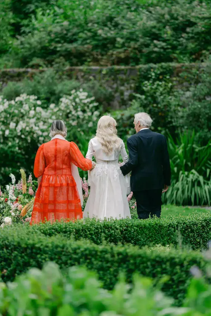 Destination wedding ceremony at Ashford Castle, County Mayo — bride linking arms with her parents before a bridal procession through lush greenery during an outdoor garden ceremony.