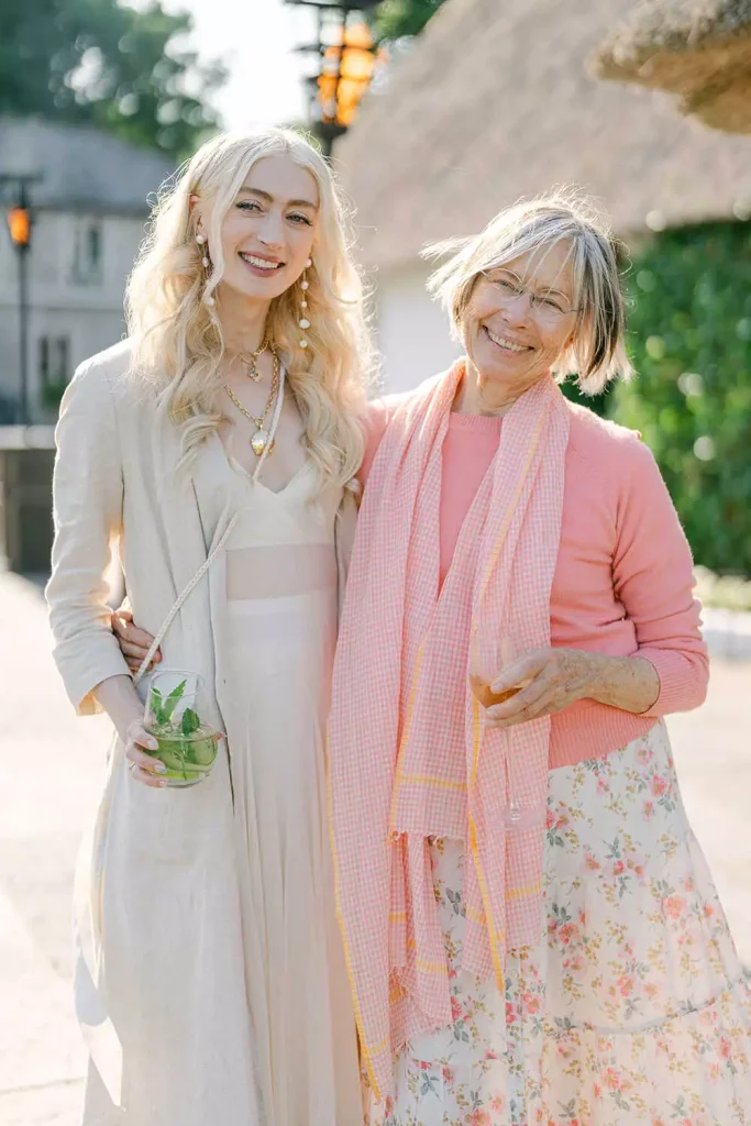 Two women smiling and posing together outdoors, one in a light beige dress holding a minty drink, the other in a pink sweater with a floral skirt and scarf.