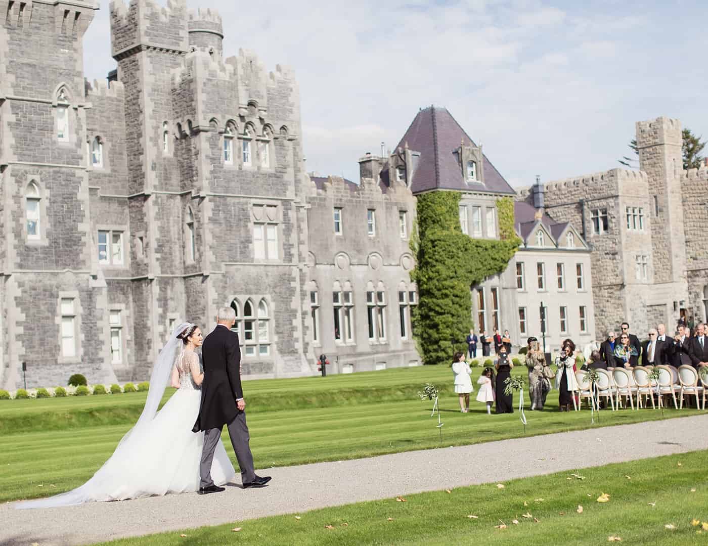 Destination wedding ceremony at Ashford Castle, County Mayo — bride and groom walking towards an outdoor ceremony on the castle lawns with guests seated against the historic estate backdrop.