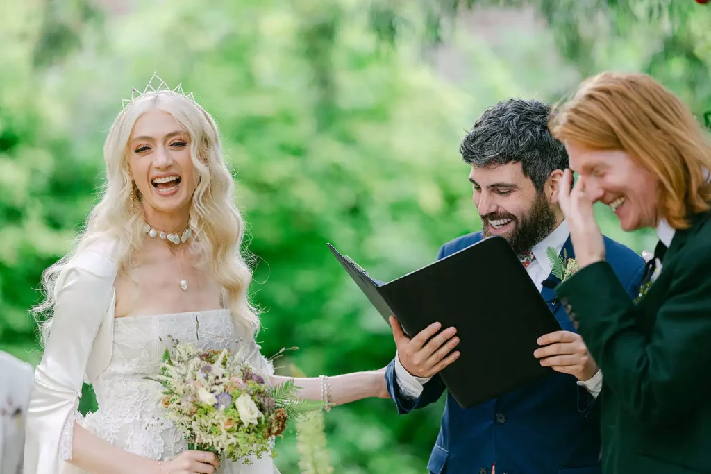 Destination wedding at Ashford Castle, County Mayo — bride and groom smiling with their celebrant holding a folder during an outdoor wedding ceremony.