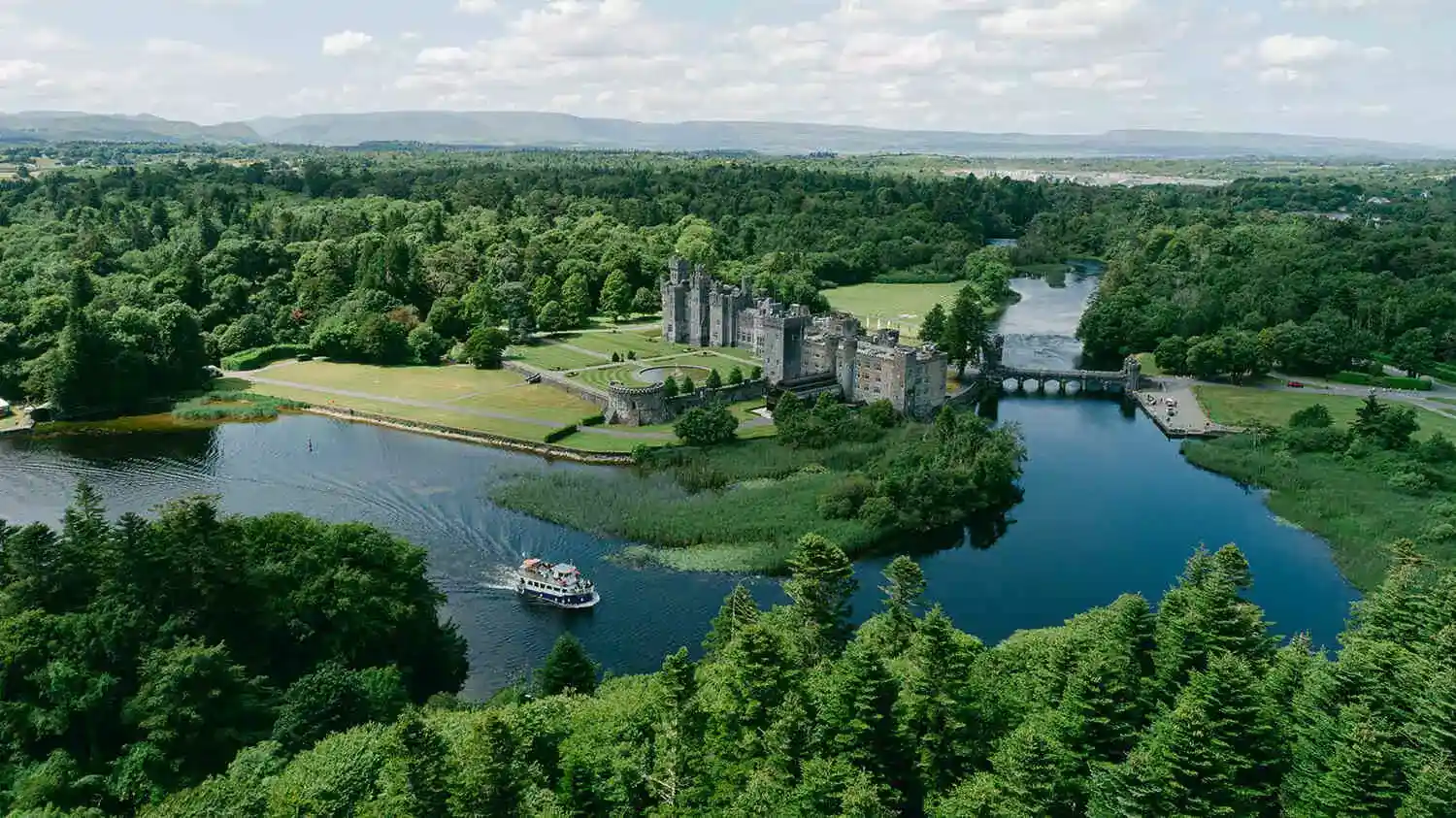 Destination wedding at Ashford Castle, County Mayo — aerial drone view of the estate showing the castle, surrounding forest, and nearby lake within the wider grounds.