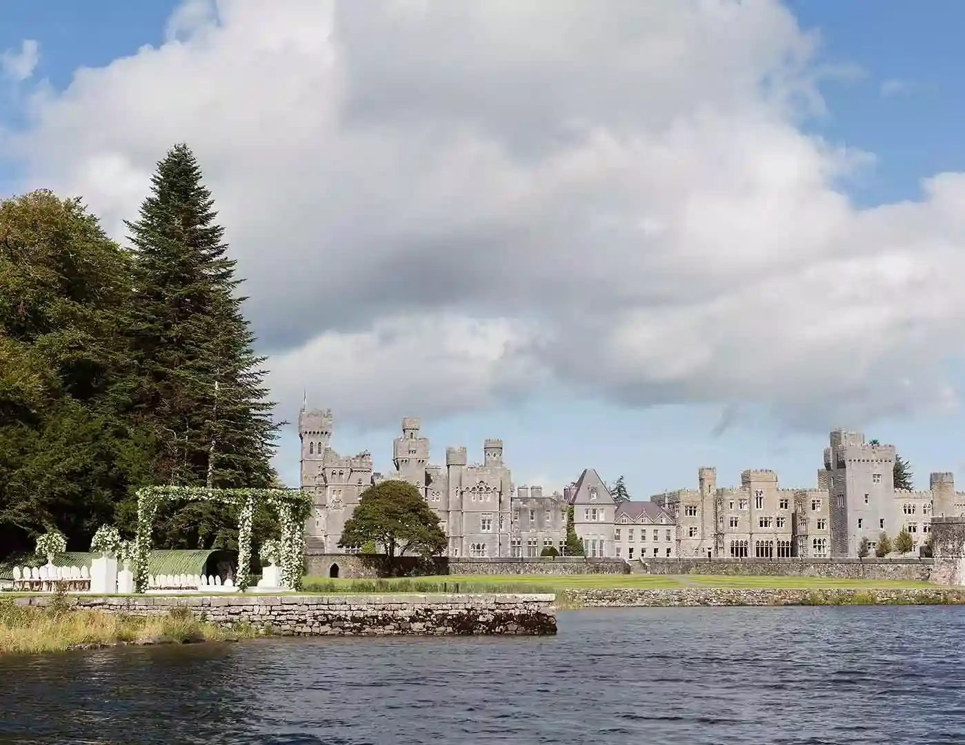 Destination wedding ceremony at Ashford Castle, County Mayo — floral arch installation set beside the lake with Ashford Castle visible in the background.