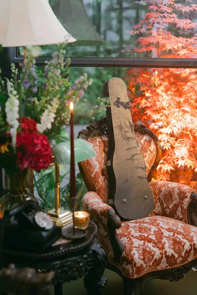 Vintage ornate chair with red damask upholstery, a guitar case resting against its back, surrounded by flowers and candlelight.