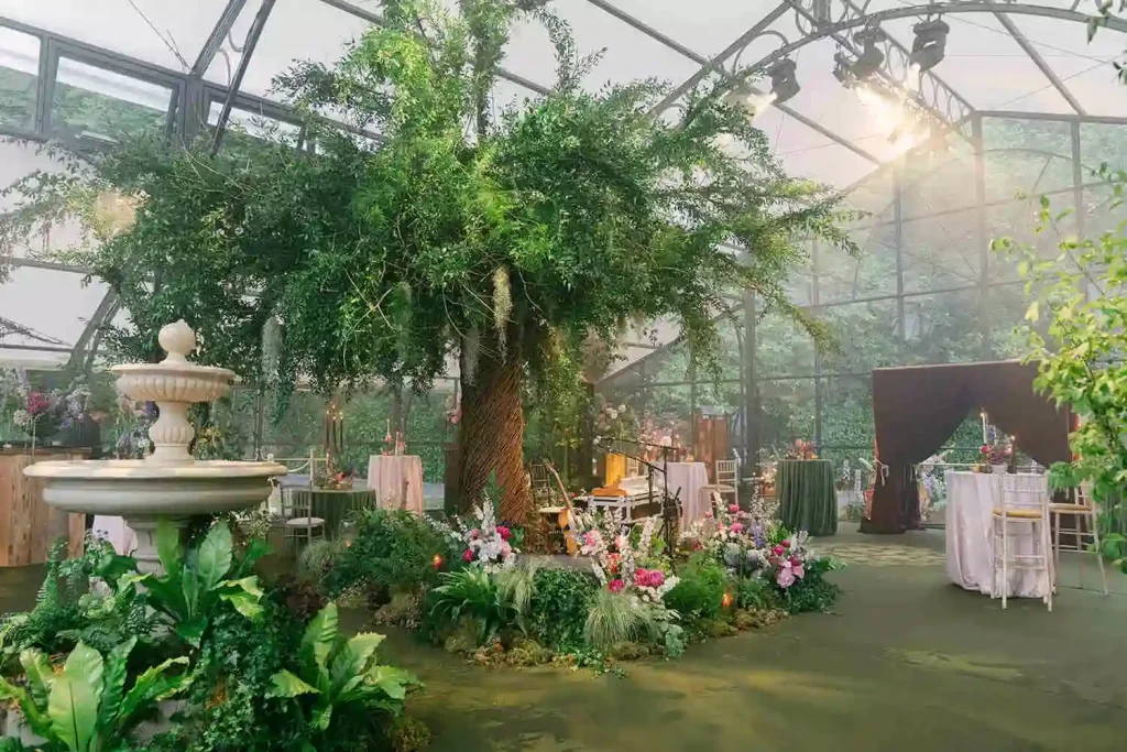 Destination wedding in Ireland at Ashford Castle, County Mayo — greenhouse orangery wedding space featuring a large willow tree installation at the centre with floral and greenery details, and a fountain installation surrounded by foliage.