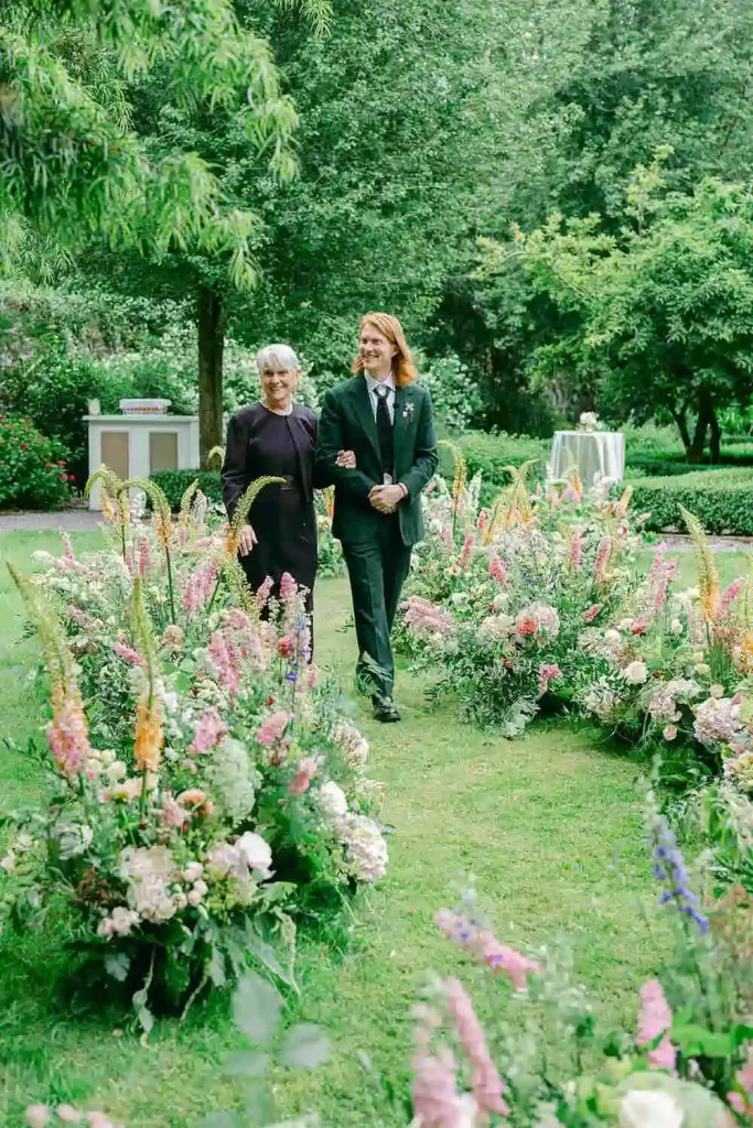 Destination wedding ceremony at Ashford Castle, County Mayo — groom walking with his mother down a curved floral aisle during an outdoor ceremony beneath the estate trees.