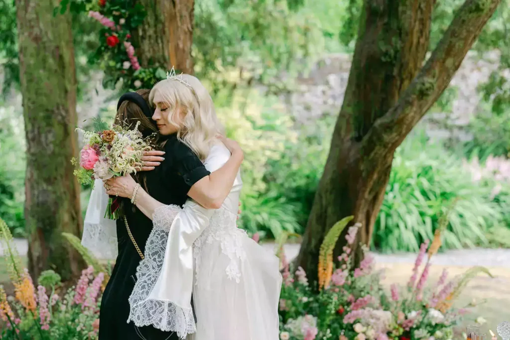 Destination wedding ceremony at Ashford Castle, County Mayo — bride embracing her wedding planner at the end of the outdoor ceremony with floral arrangements visible in the background.