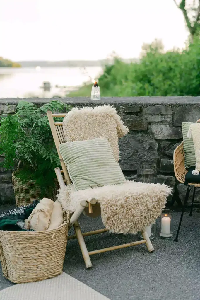 Destination wedding in Ireland at Ashford Castle, County Mayo — lounge seating detail featuring a wooden chair with a fur throw and cushion, blankets in a basket, a leafy green plant, and candles arranged on the ground.