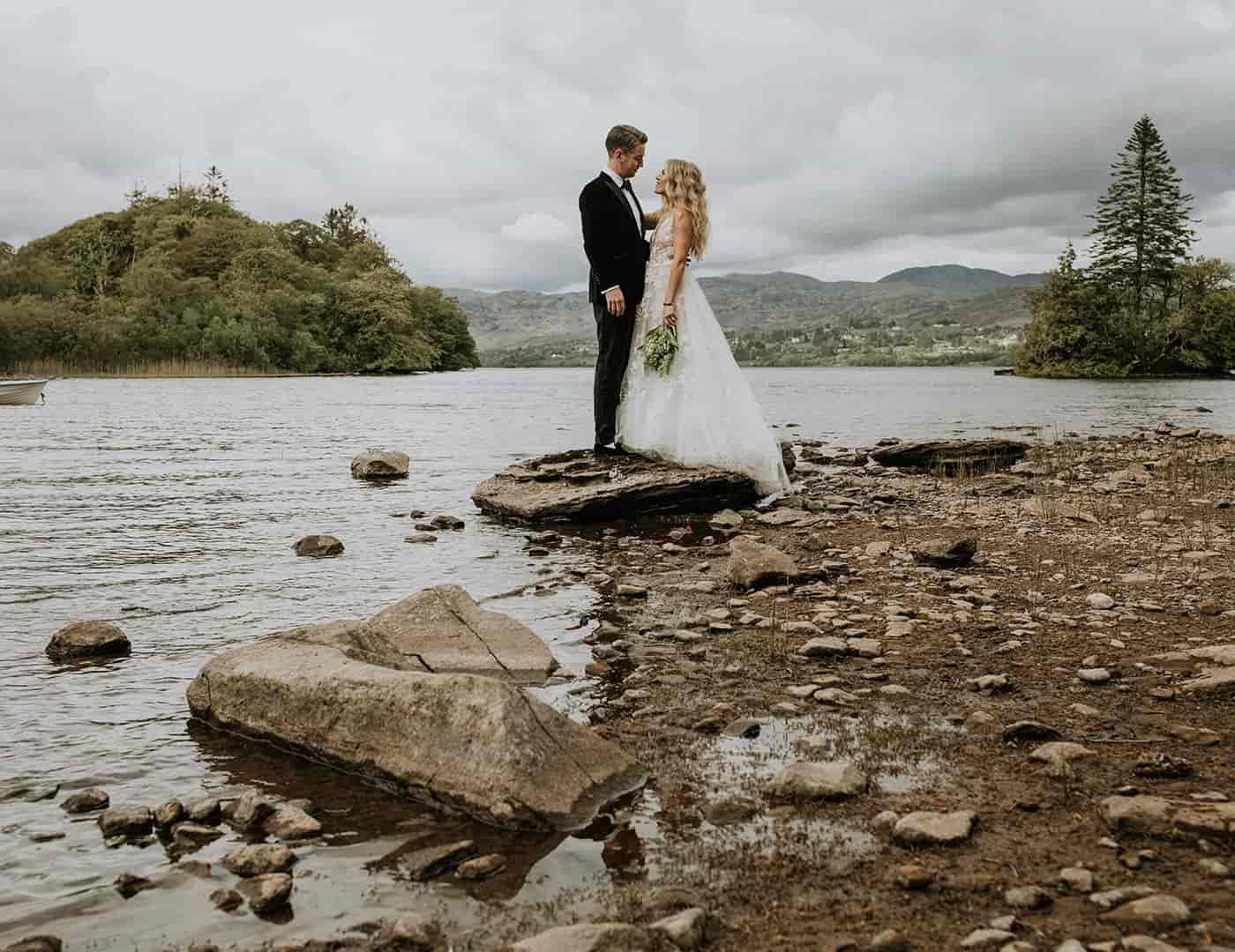 Destination wedding in Ireland — bride and groom embracing outdoors beside a lake landscape with mountains in the background.