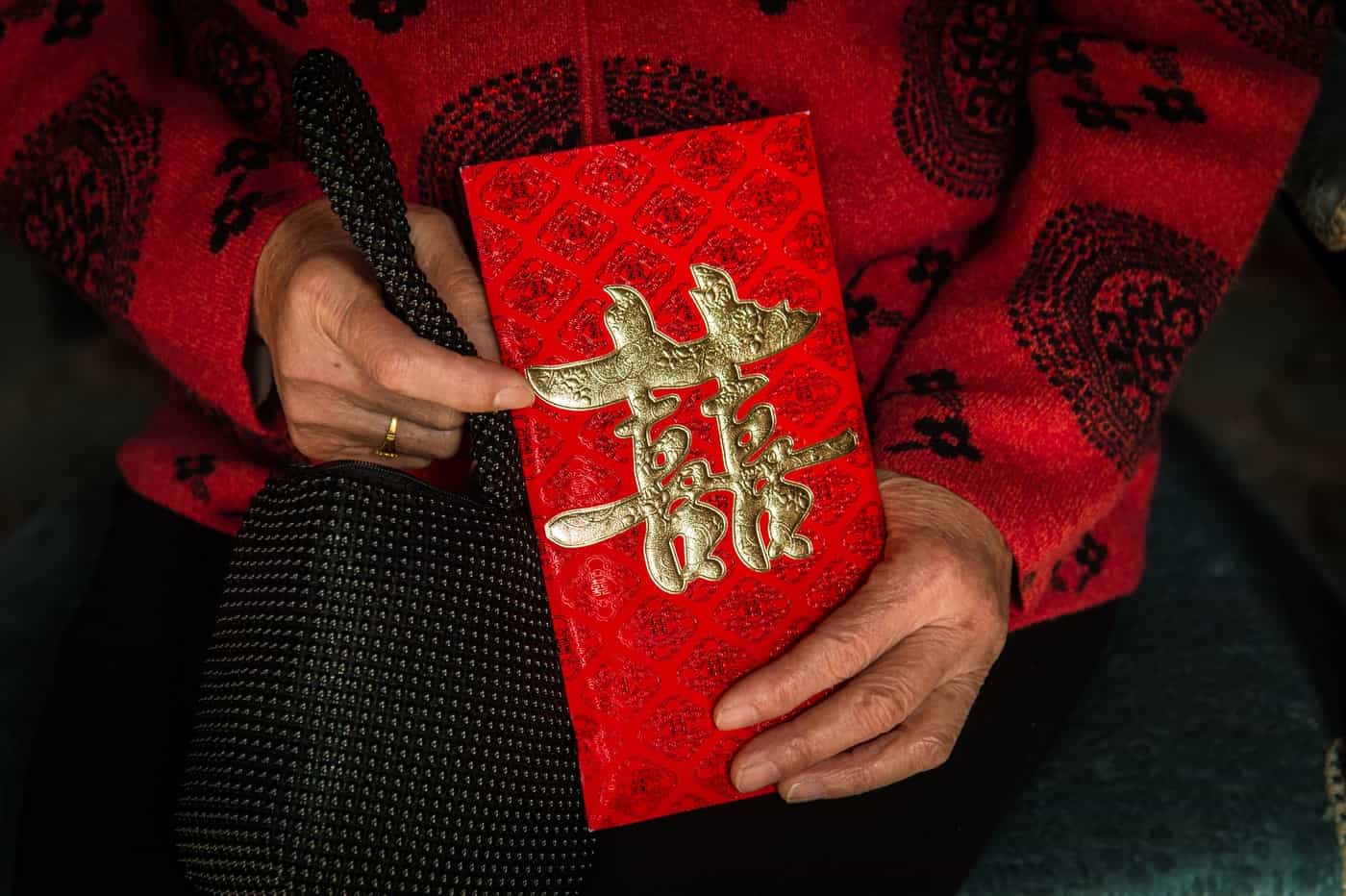 Elderly person in a red patterned sweater holding a red Chinese New Year envelope with gold characters.