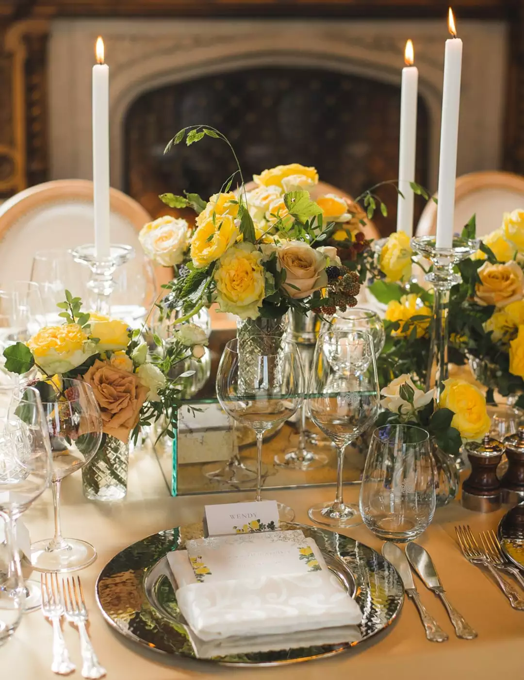 Elegant dining table with yellow and cream floral centerpieces, tall white candles, and crystal glassware set for a formal event.
