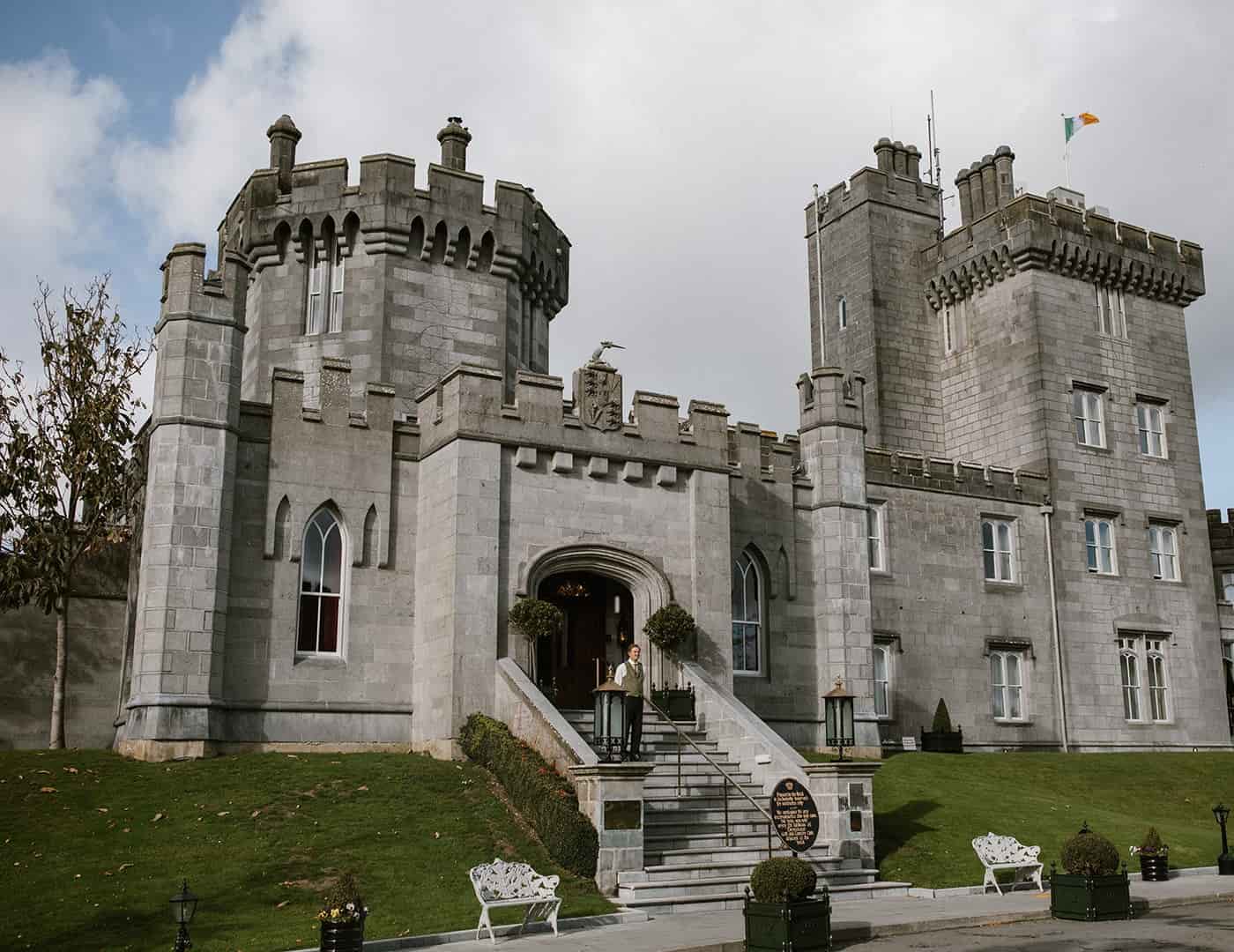 Grey stone castle-style mansion with crenellated towers, arched windows, and a person standing on the front steps.