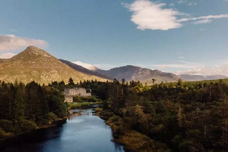 River winds through a dense forest with a white building on the left and mountains in the distance under a blue sky.