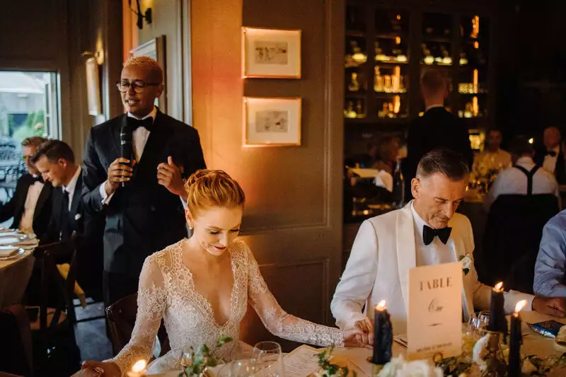 Bride in a lace wedding gown sits at a candlelit head table while a suited man, holding a microphone, gives a speech nearby; guests in formal attire mingle in the background.