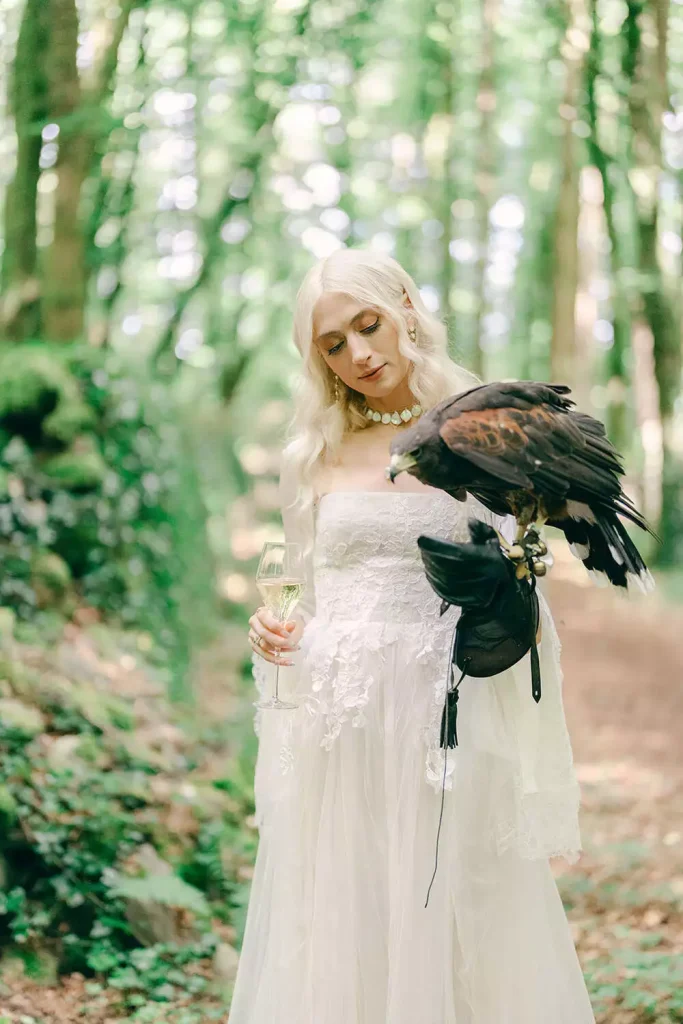 Woman in a white lace dress holding a champagne glass with a large black bird perched on her forearm in a green forest setting