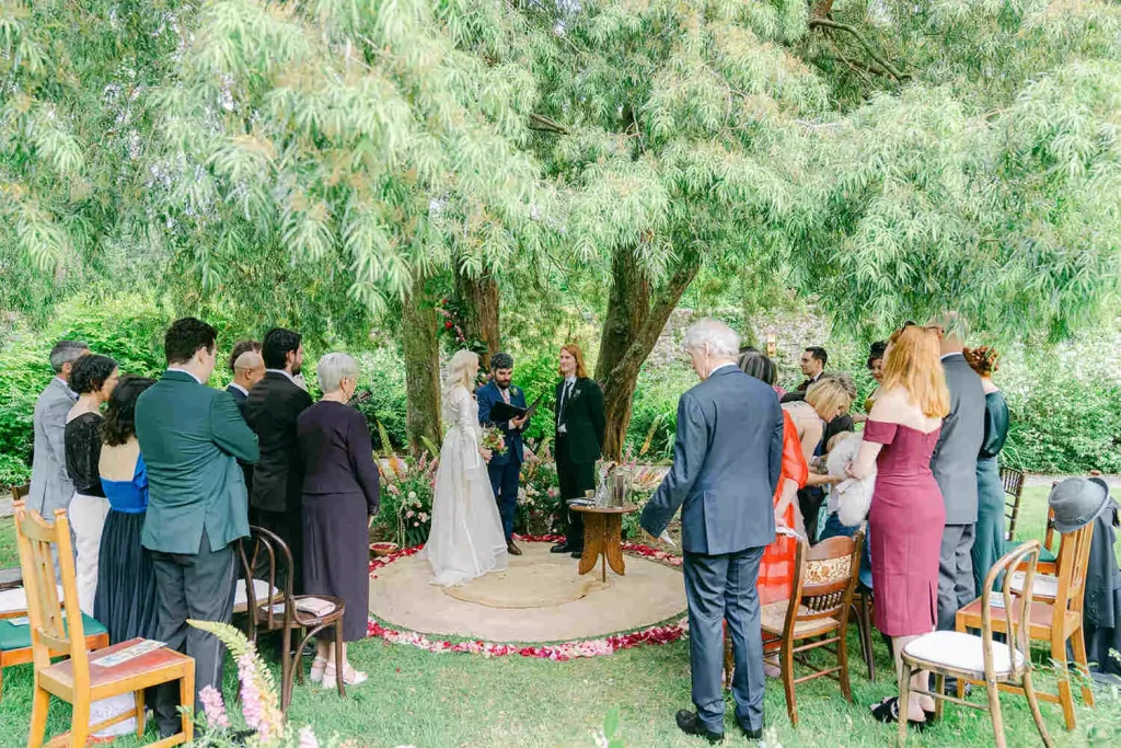 Outdoor garden wedding ceremony under a canopy of trees with guests standing around a circular platform as the couple exchanges vows.