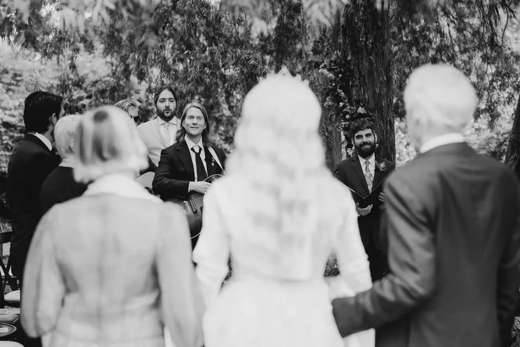 Irish destination wedding at Ashford Castle, County Mayo — bride and her parents at the end of the processional with the groom holding a guitar beside his groomsmen during an outdoor ceremony.