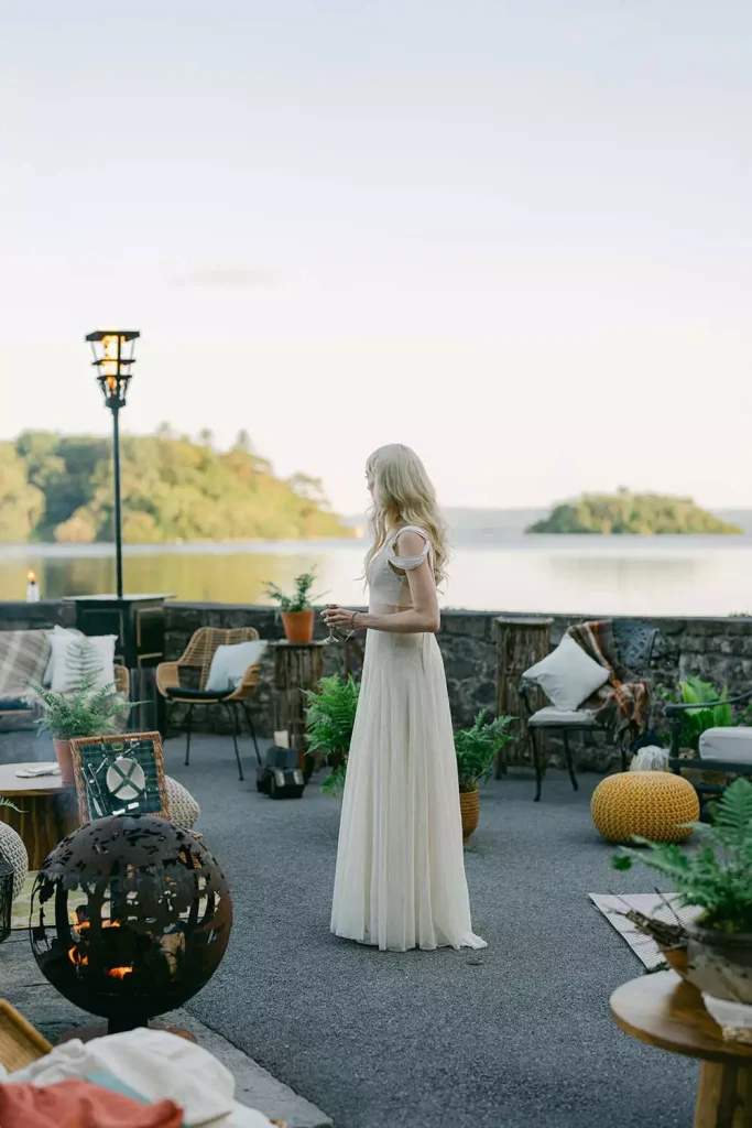 Irish destination wedding at Ashford Castle, County Mayo — bridal portrait on a lakeside dock surrounded by lounge furniture and greenery, holding a drink while looking across the water.