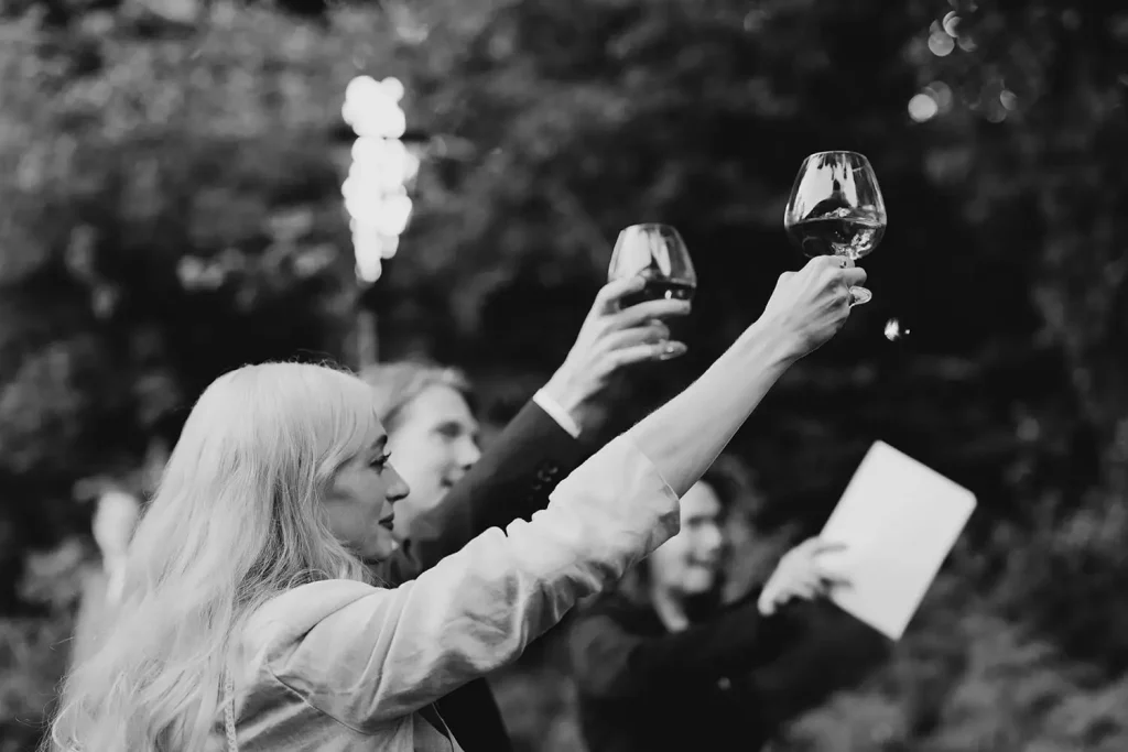 People raise wine glasses in a toast at an outdoor celebration, blurred greenery in the background (black and white).