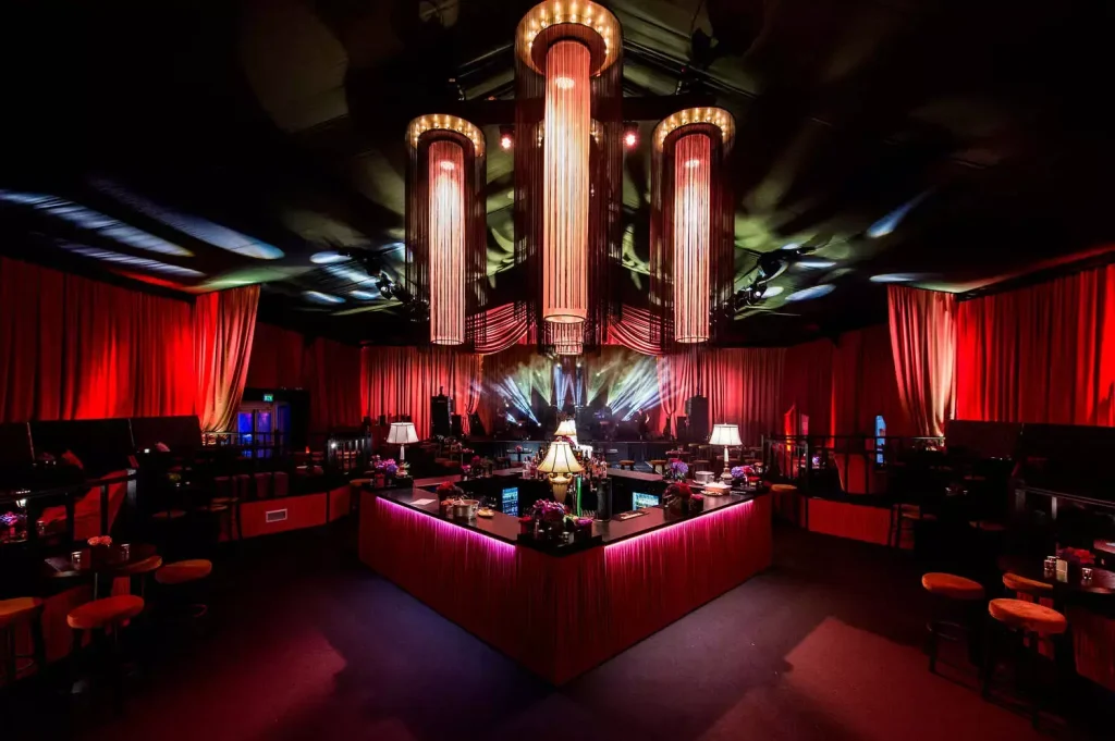 Upscale event venue with a central glowing U-shaped bar, red drapes, and dramatic cylindrical chandeliers hanging from the ceiling; stage lighting at the far end.