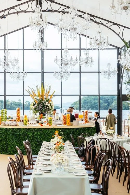 Destination wedding at Ashford Castle, County Mayo — long brunch table inside a glass marquee with chandeliers, a breakfast bar decorated with fruit, and views across Lough Corrib.