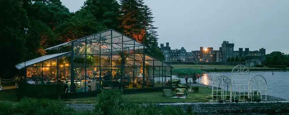 Destination wedding at Ashford Castle, County Mayo — illuminated exterior of the orangery at night with the castle visible in the background across the estate grounds.