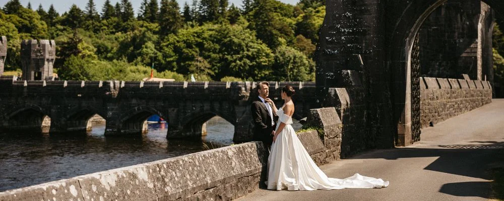 Destination wedding at Ashford Castle, County Mayo — bride adjusts the groom's tie during a portrait on the estate grounds, with the river visible in the background on a sunny day.
