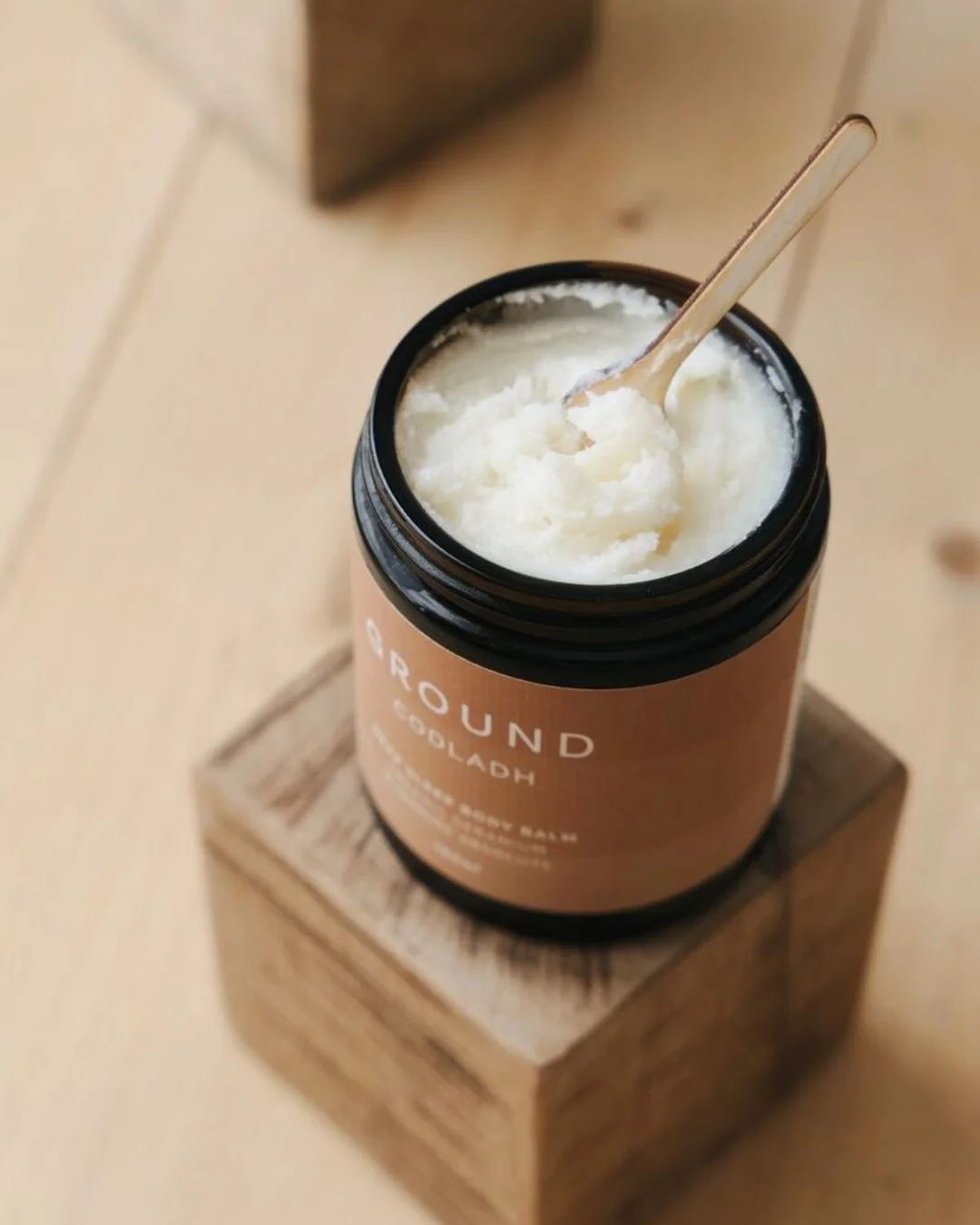 Open jar of white creamy balm or lotion with a wooden spoon resting inside, placed on a wooden block. (Skincare product)