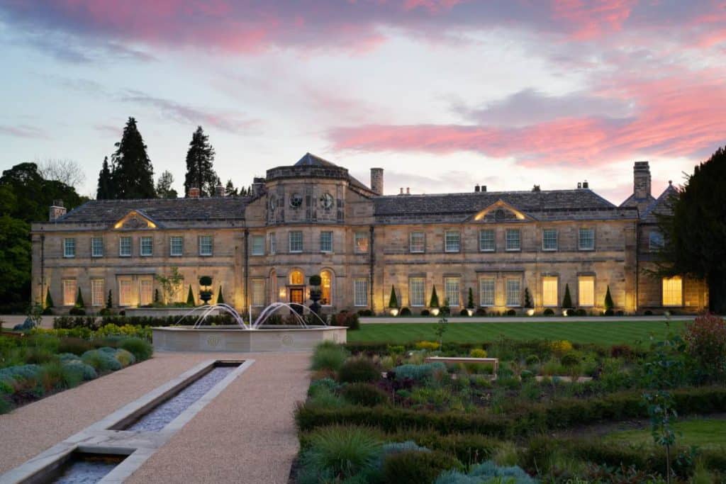 Grand stone manor with lit windows and a central fountain, set in formal gardens at dusk.