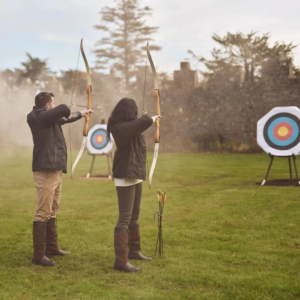 Irish castle wedding at Adare Manor, County Limerick — outdoor archery activity with two participants holding bows and arrows aimed toward distant targets set across the estate grounds within a softly misted landscape.