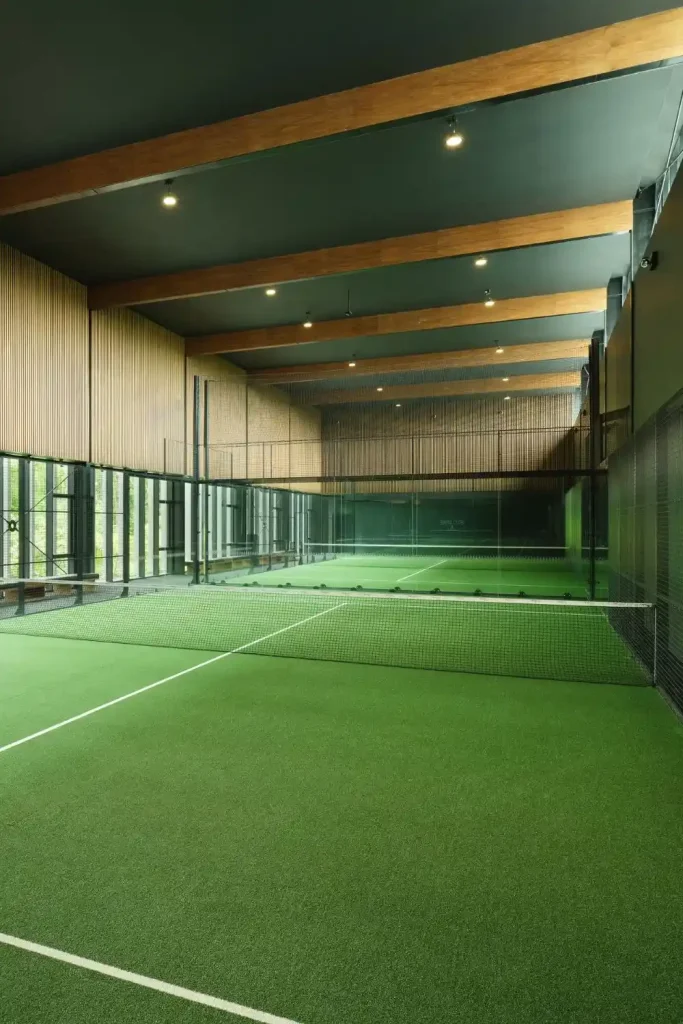 Indoor tennis court with green turf, net across center, glass walls, and wooden ceiling beams.