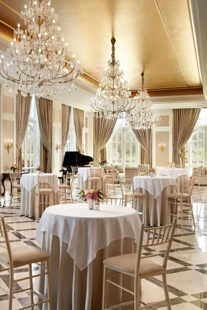 Elegant banquet hall with crystal chandeliers, round tables in white linens, and beige chairs under draped windows.