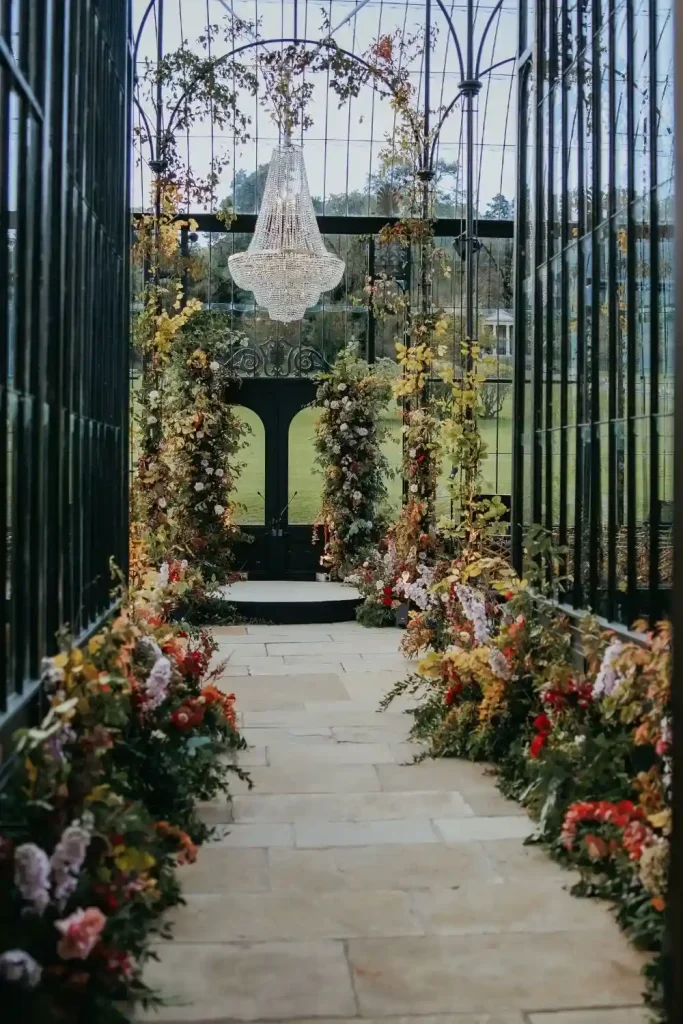 Destination wedding in Ireland Ballyfin Demesne, County Laois — The Orangery arranged for a ceremony with a floral aisle in autumn tones, leading to a large floral installation beneath a statement chandelier suspended from the ceiling.