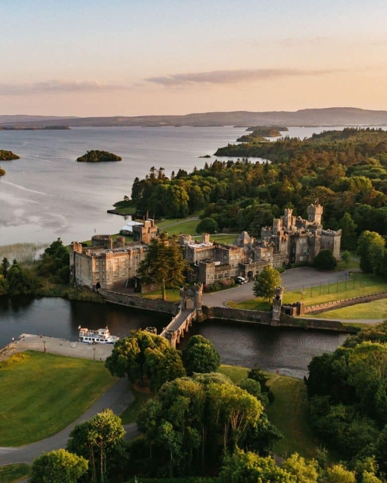 A historic castle with a stone bridge spanning a moat, surrounded by trees and green lawns beside a calm lake at sunset.