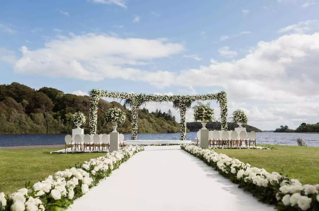 Outdoor lakeside wedding setup with a white flower-covered aisle and arched floral arch, rows of white chairs on either side.