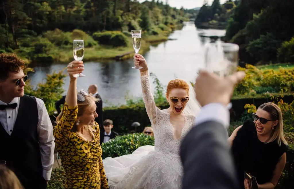 Bride in lace dress toasts with champagne as friends cheers outdoors by a river backdrop and fountains.