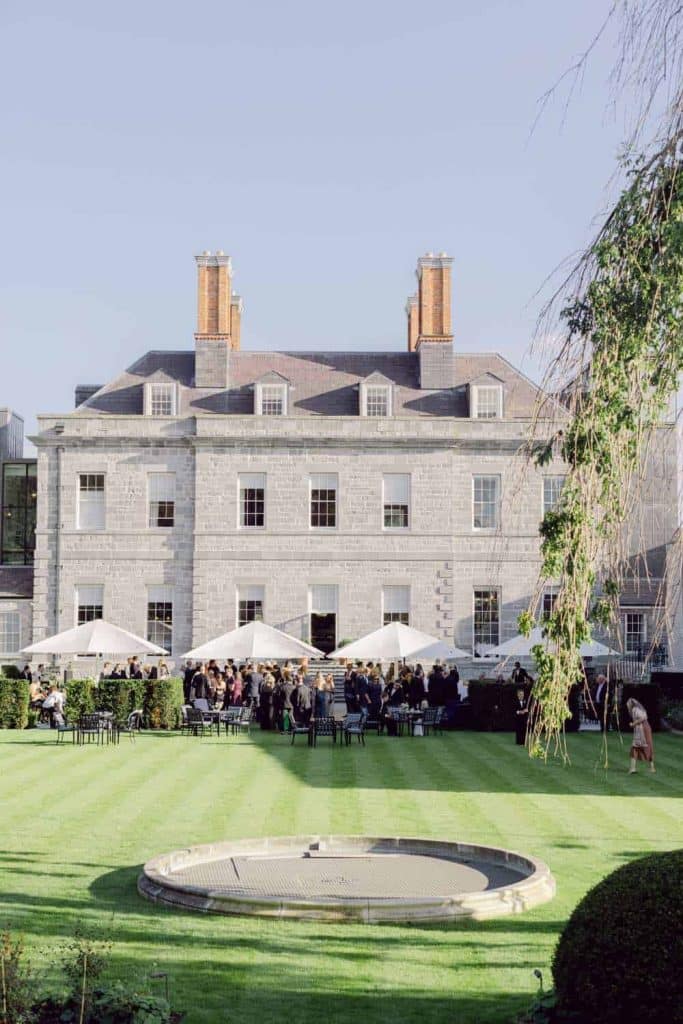 Destination wedding ceremony at Cashel Palace, County Tipperary — bright exterior drinks reception on the estate grounds with guests mingling beneath white parasols during an outdoor wedding gathering.