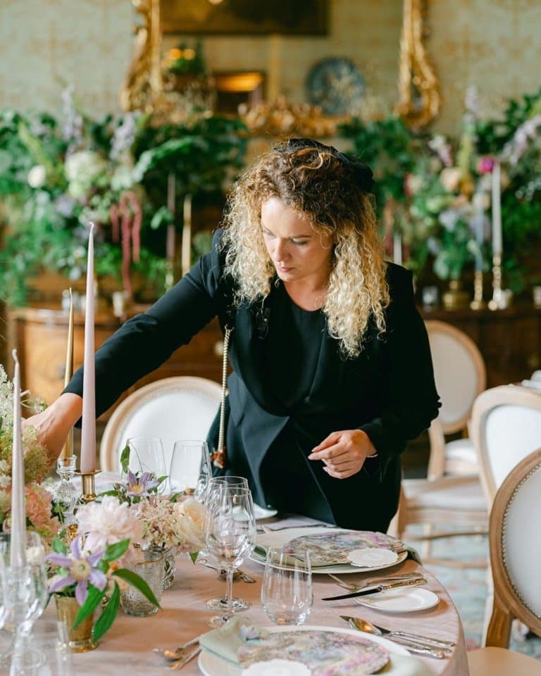 Woman in a black blazer arranging a formal dining table with candles and floral centerpiece in an elegant room.